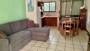 A living room with a grey sofa, a dining table with chairs, a kitchen area, and a window with curtains. - 2bdrm/2bth walk to the beach1 (Coco)
