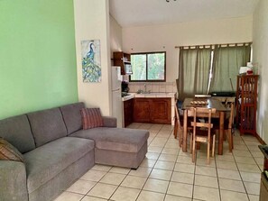 A living room with a sofa, a dining table with chairs, a kitchen area, and a window with curtains.