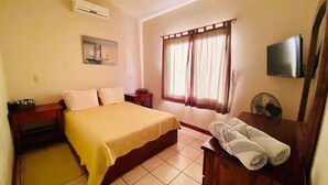 A hotel room with a bed, a bedside table, a TV, a window with curtains, and a wooden dresser with towels. - 2bdrm/2bth walk to the beach1 (Coco)