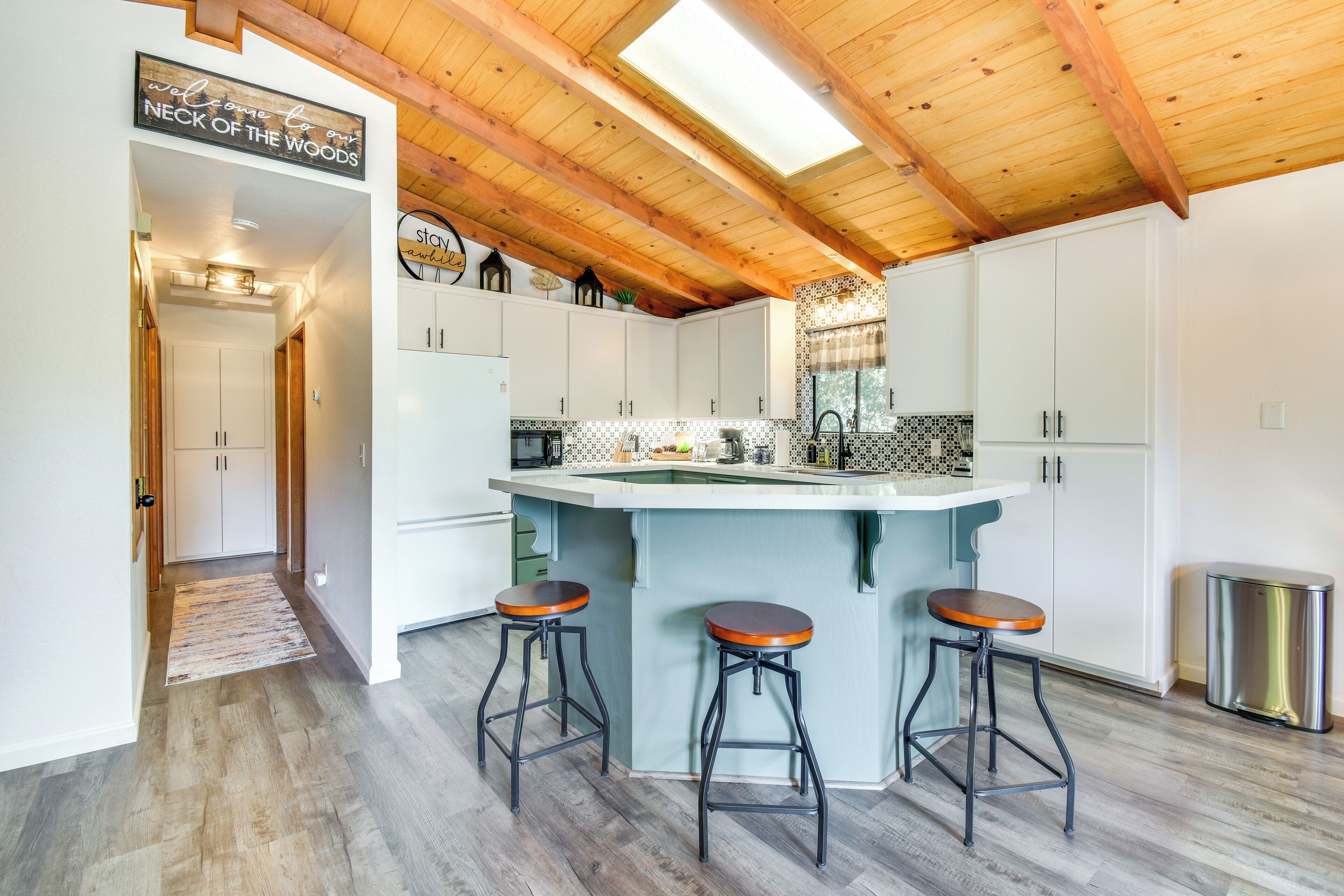 Une cuisine avec un plafond en bois, des armoires blanches, un îlot central avec des tabourets et une pancarte sur laquelle on peut lire : « Bienvenue à Neck of the Woods ».