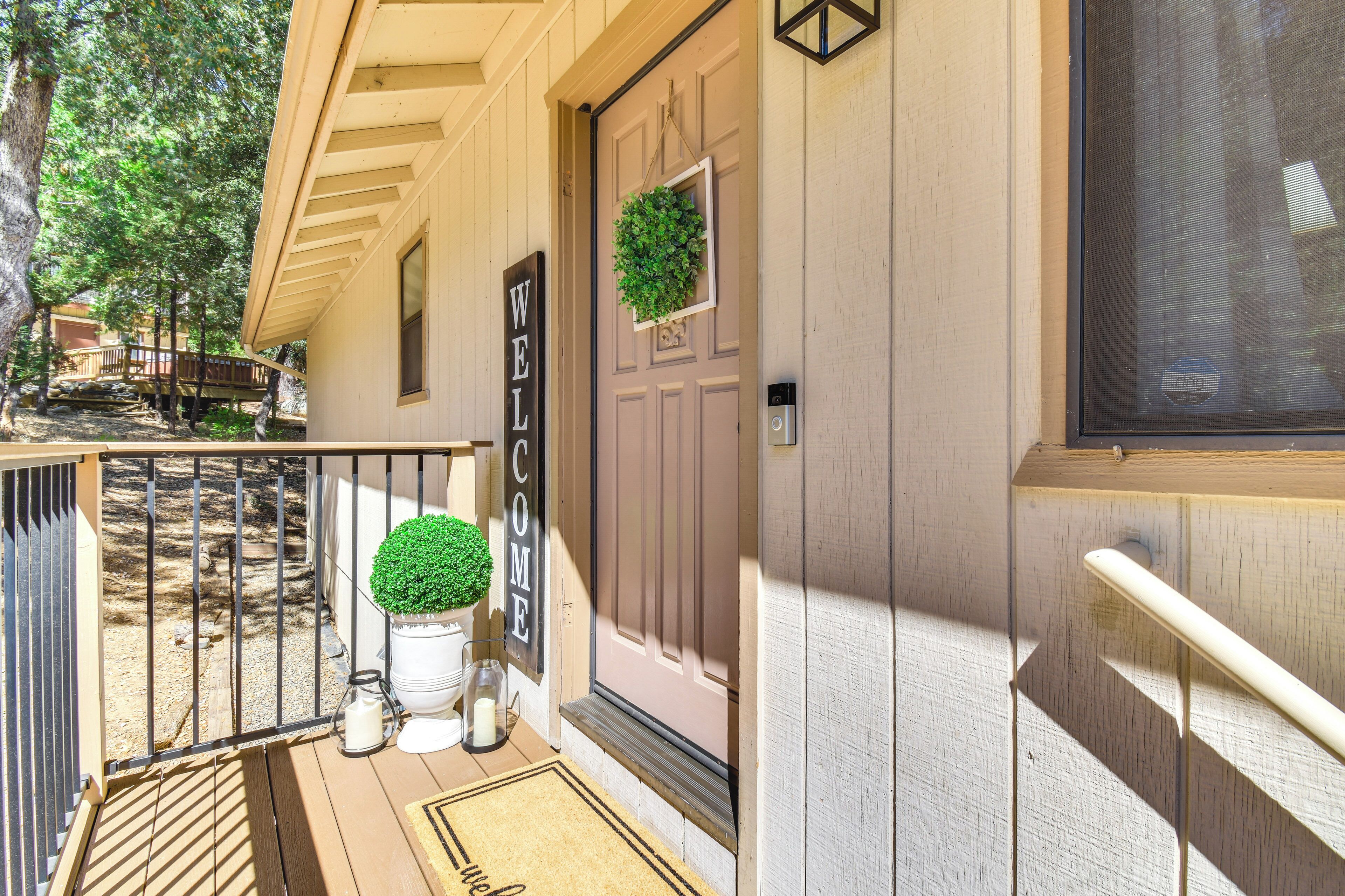 Une maison en bois avec une pancarte « BIENVENUE », une plante en pot et un carillon.