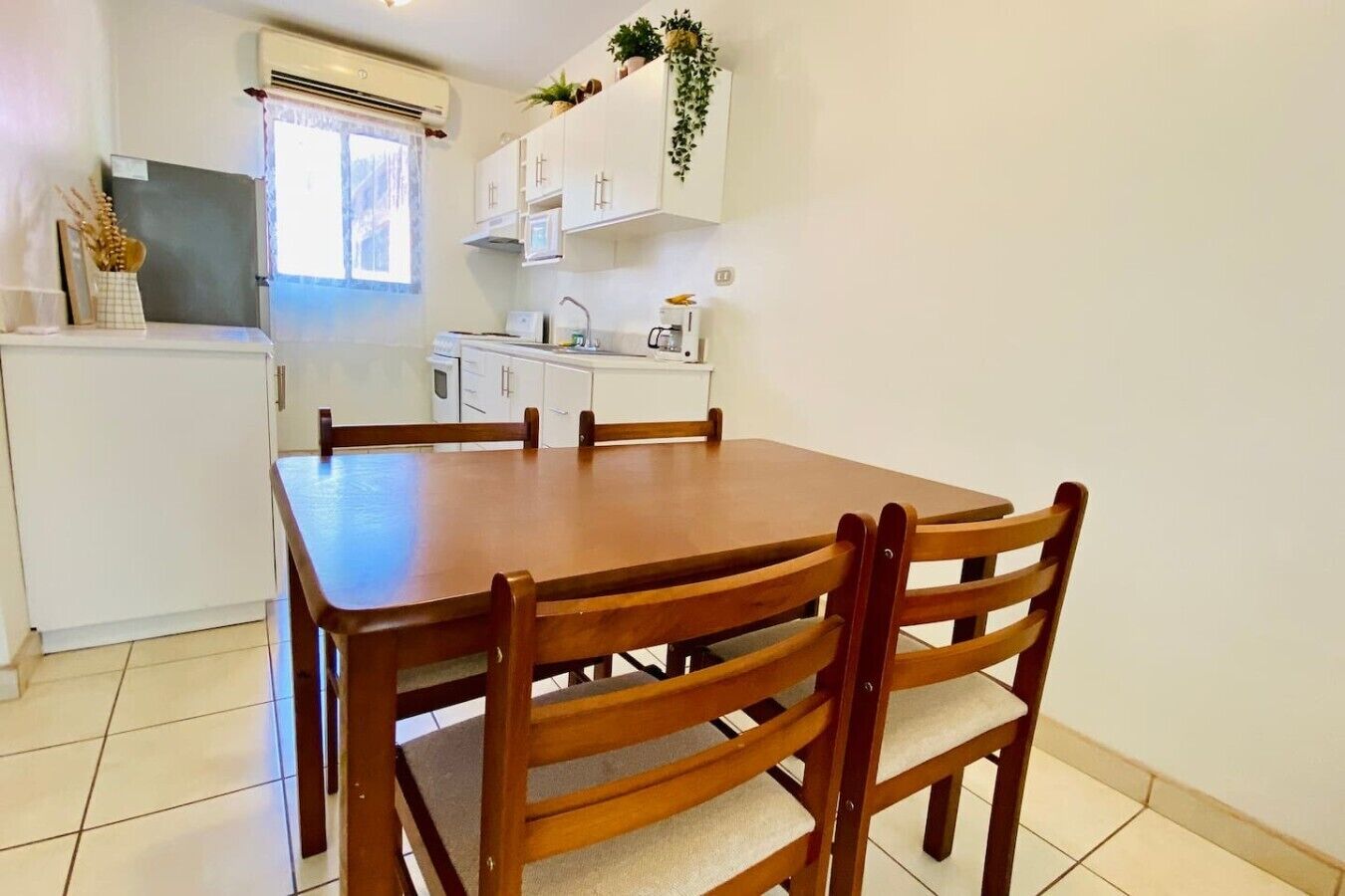A dining area with a wooden table and chairs, a kitchen in the background, and a white wall with a plant.