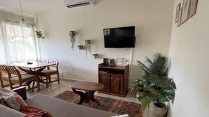 Ein Wohnzimmer mit einer Couch, einem Esstisch mit Stühlen, einem an der Wand befestigten Fernseher und mehreren Blumentöpfen.