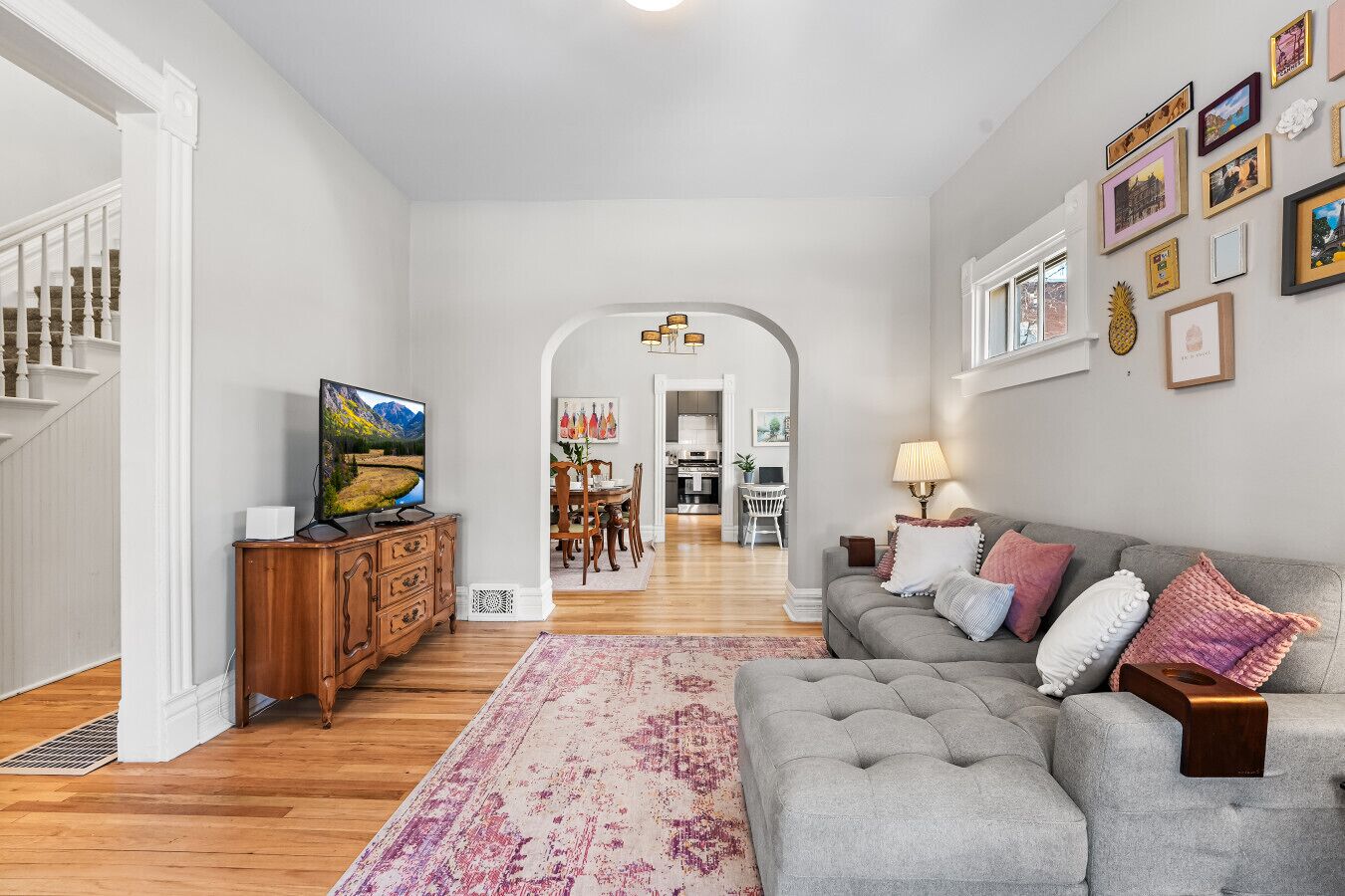 A spacious living room with a grey sofa, a wooden TV stand, and a dining area in the background.