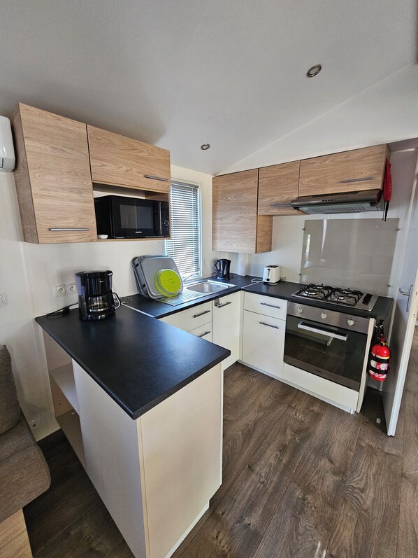 A compact kitchen with wooden cabinets, a microwave, a kettle, a coffee maker, a gas stove, and a fire extinguisher.