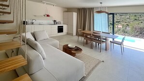 A modern living room with a sofa, dining table, and chairs. There is a kitchen area with cabinets and a window. - Luxurious home in Zman (Zman)