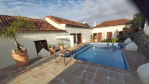 A swimming pool with a tiled deck, surrounded by terracotta-roofed buildings, potted plants, and a stone wall.