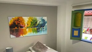 Een slaapkamer met een bed, nachtkastje, lamp, schilderij en een raam met uitzicht.