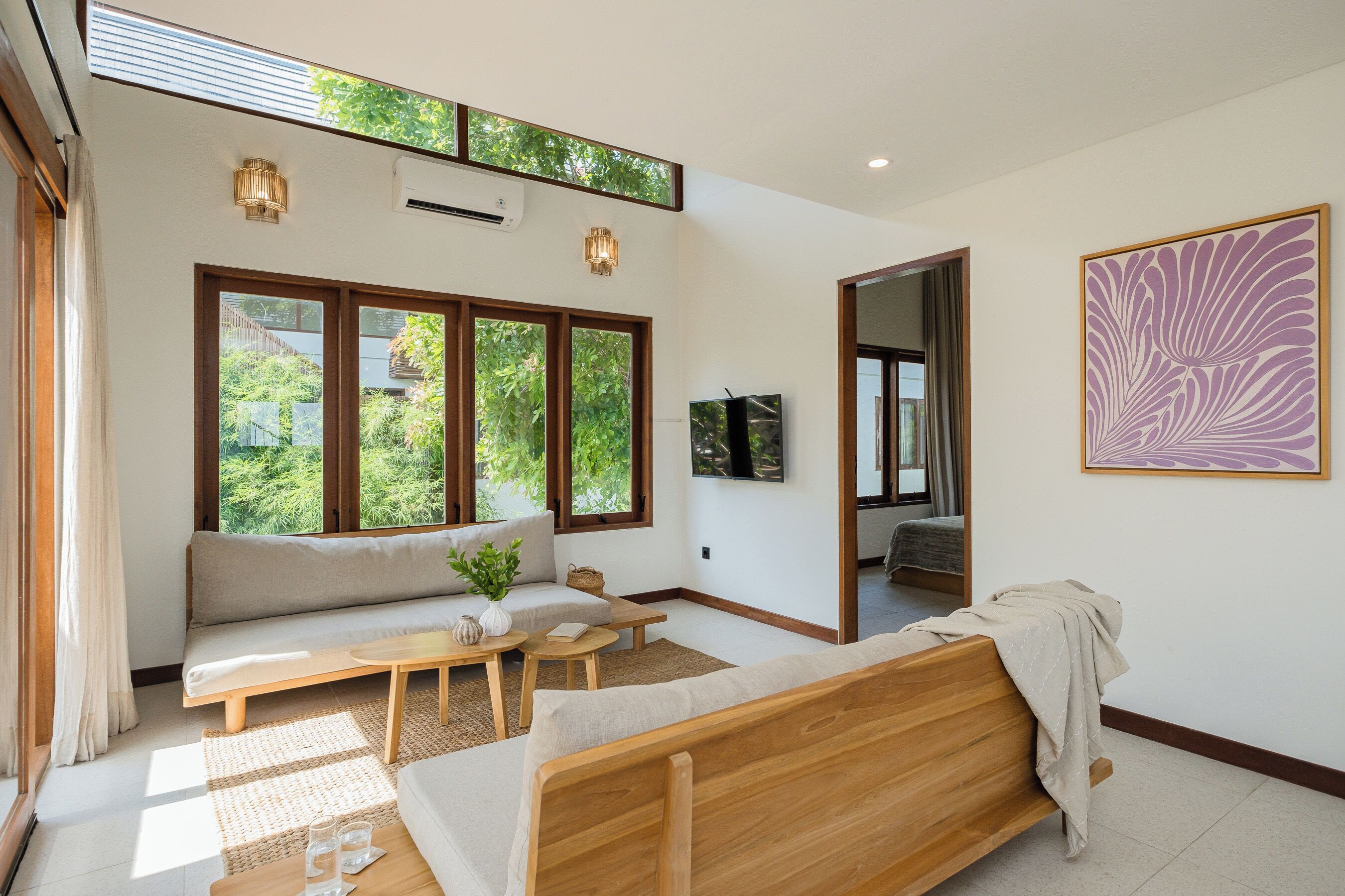 A modern room with a sofa, a wooden bed, a TV, and large windows offering a view of greenery.