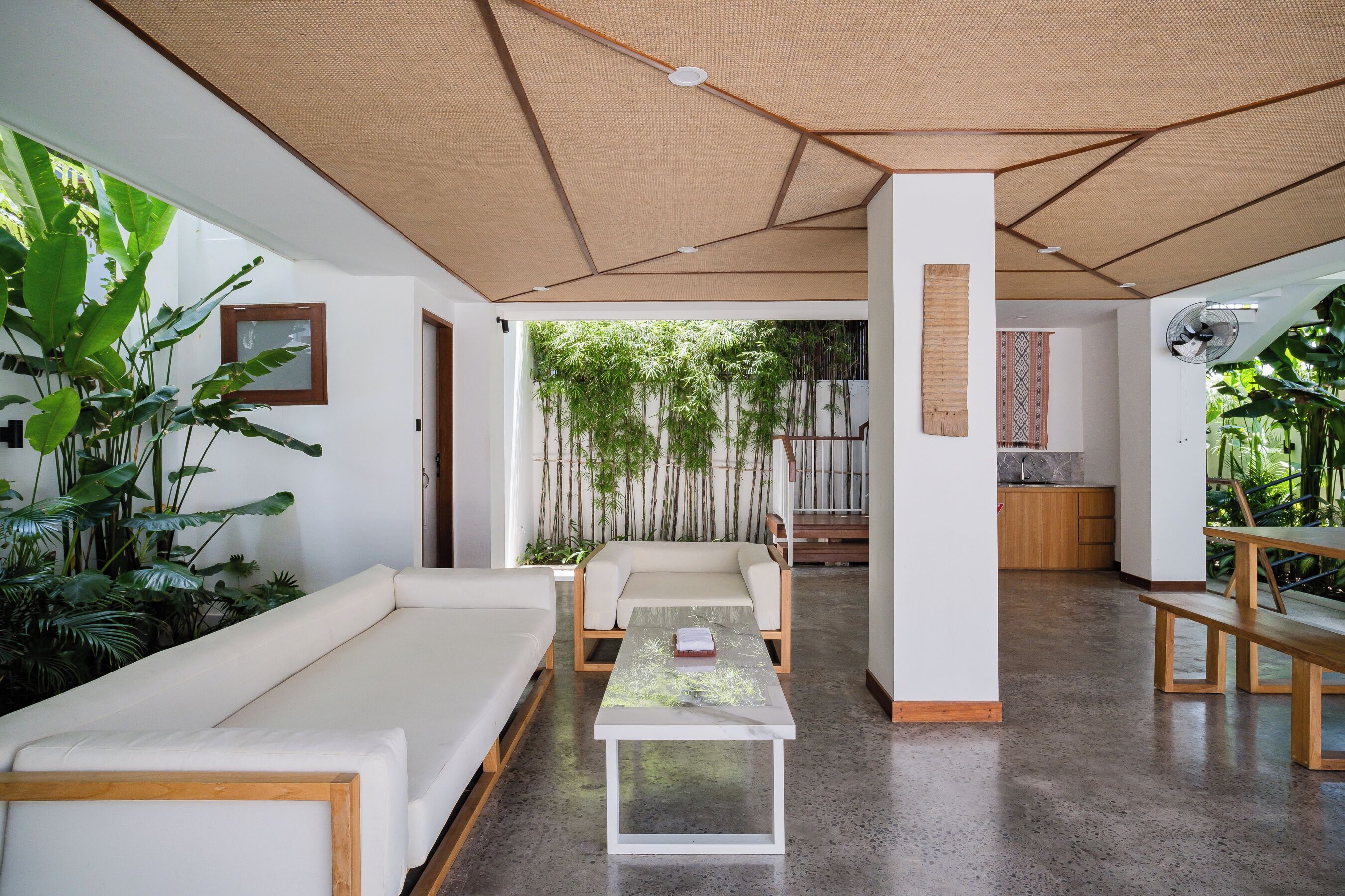 A modern interior with a white sofa, a wooden bench, and a glass-top coffee table. There is a bamboo ceiling and a view of a garden with plants and a wooden structure.