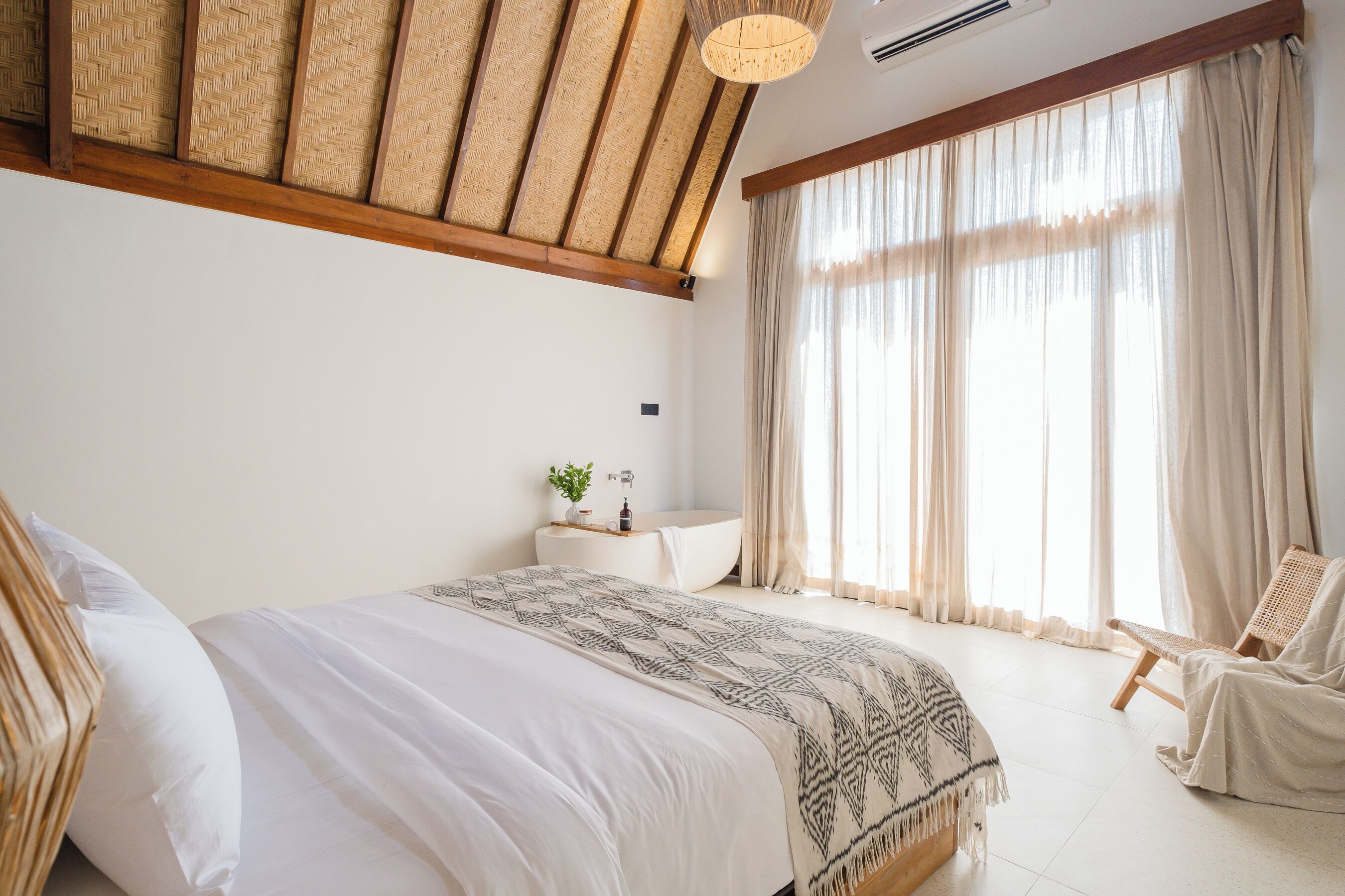 A bedroom with a bed, a bedside table with a plant, a window with curtains, and a chair.
