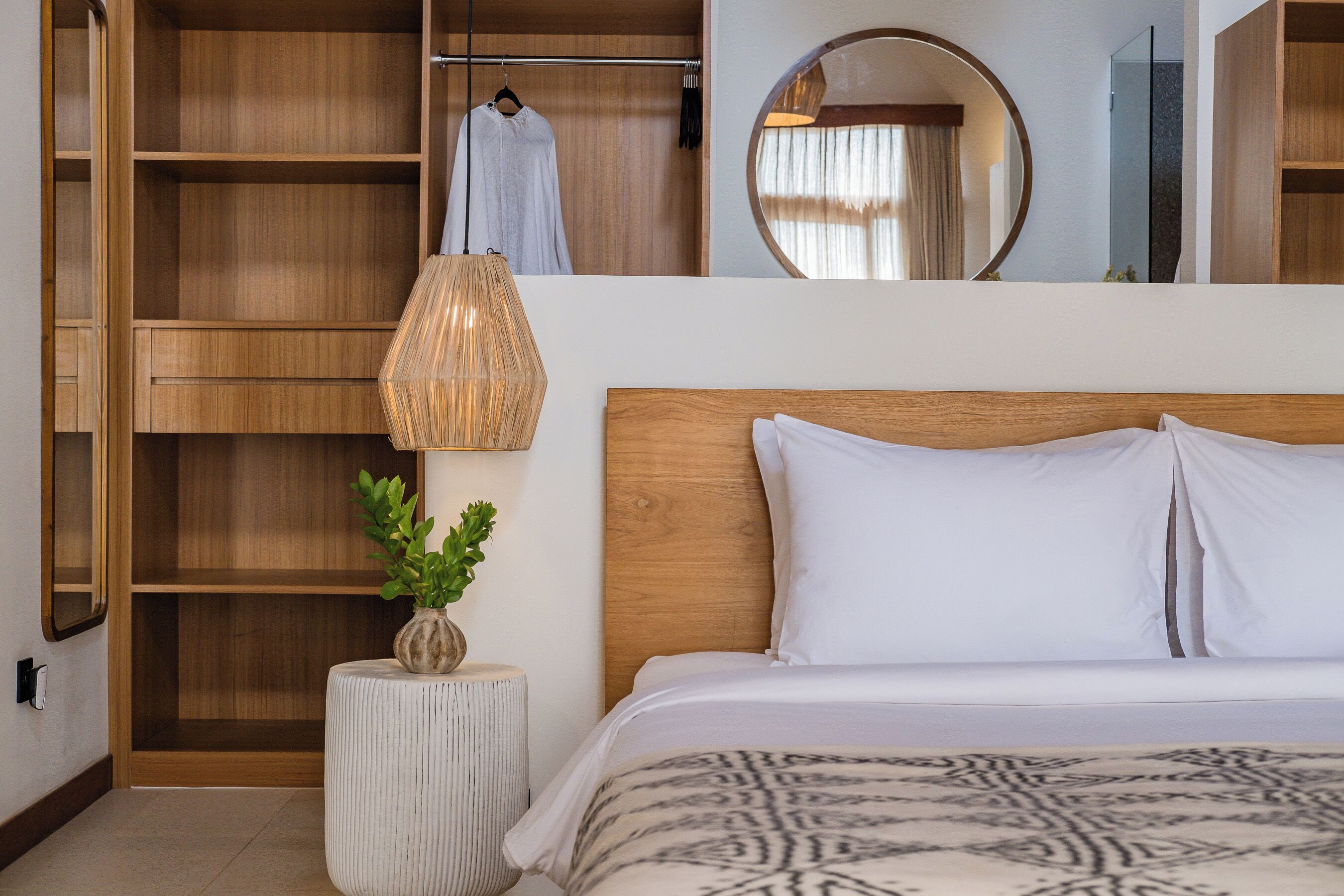 A bedroom with a wooden headboard, a bed with white bedding, a round mirror, a hanging light fixture, and a wardrobe with open doors.