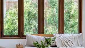 A wooden bed with a white pillow and a plant in a vase, a woven basket, and a view of green foliage through large windows. - Treetop Villas (Pecatu)