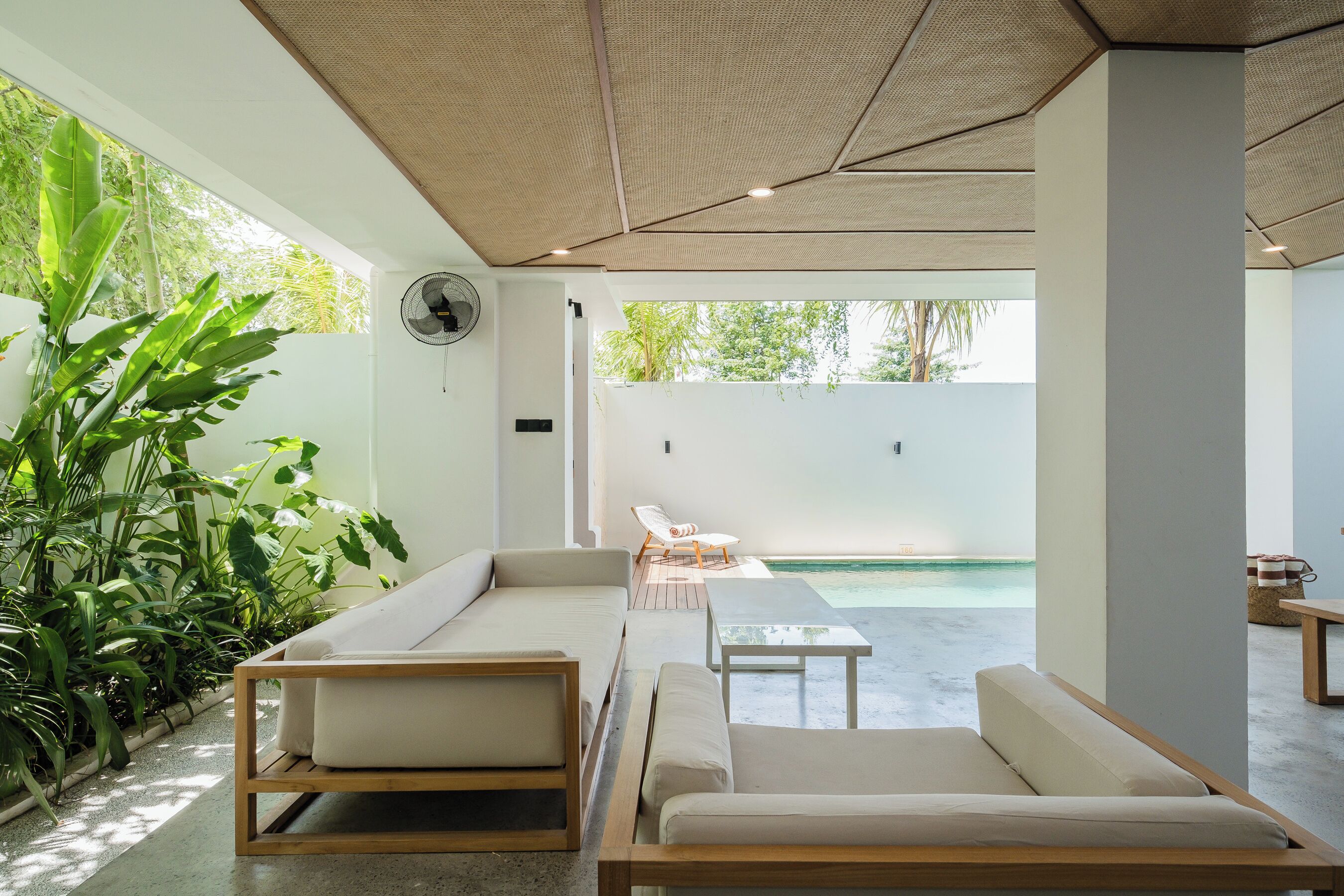 A modern outdoor seating area with sofas and a table, surrounded by lush greenery and a view of a pool.