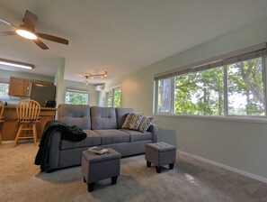 A modern living room with a gray sofa, two ottomans, a ceiling fan, and a view of trees through large windows.