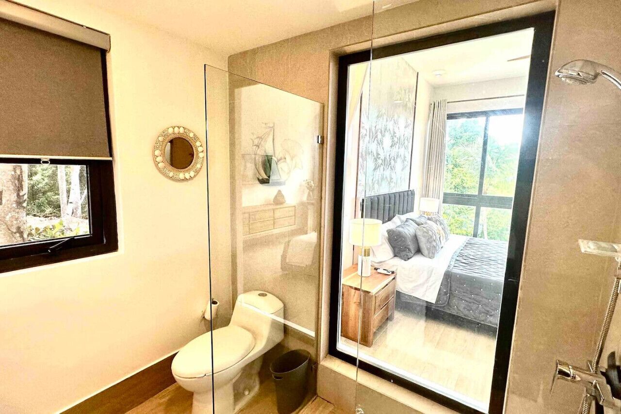 A modern bathroom with a toilet, a glass-enclosed shower, and a large window offering a view of a bedroom with a bed and curtains.