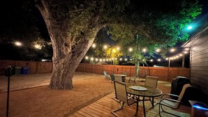 Ein Garten mit Holzterrasse, einem großen Baum, Gartenmöbeln und Lichterketten.