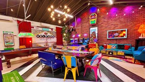 Ein lebhafter Spielsalon mit Billardtisch, bunten Sesseln, Neonreklamen und einer Ziegelwand im Hintergrund.