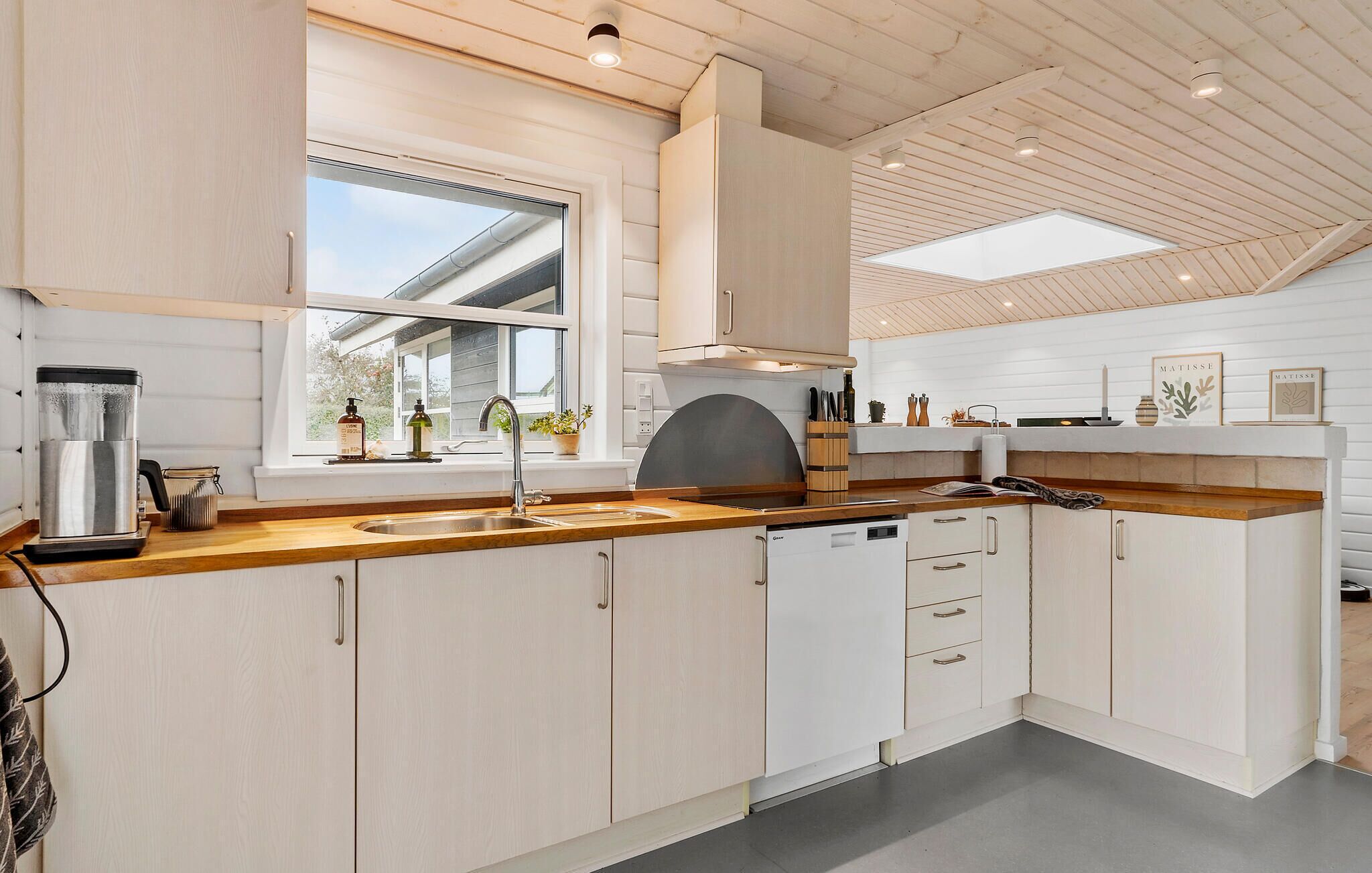 Une cuisine moderne avec des meubles blancs, un plan de travail en bois, un lave-vaisselle encastré et une fenêtre qui laisse entrer la lumière naturelle.
