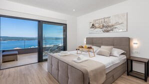 Une chambre avec un grand lit, une table de chevet avec une lampe, un tableau et une vue sur la mer par une porte vitrée.