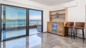Une chambre moderne dotée d’une porte vitrée donnant sur un balcon avec vue sur la mer, d’un meuble en bois contenant de la verrerie et de deux fauteuils rembourrés.