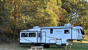 A white RV with a slide-out dining area and a small table, parked in a grassy field with trees in the background. - Horn and Thistle Farm - Camper/RV in Abbottstown, Pennsylvania, United States (Abbottstown)