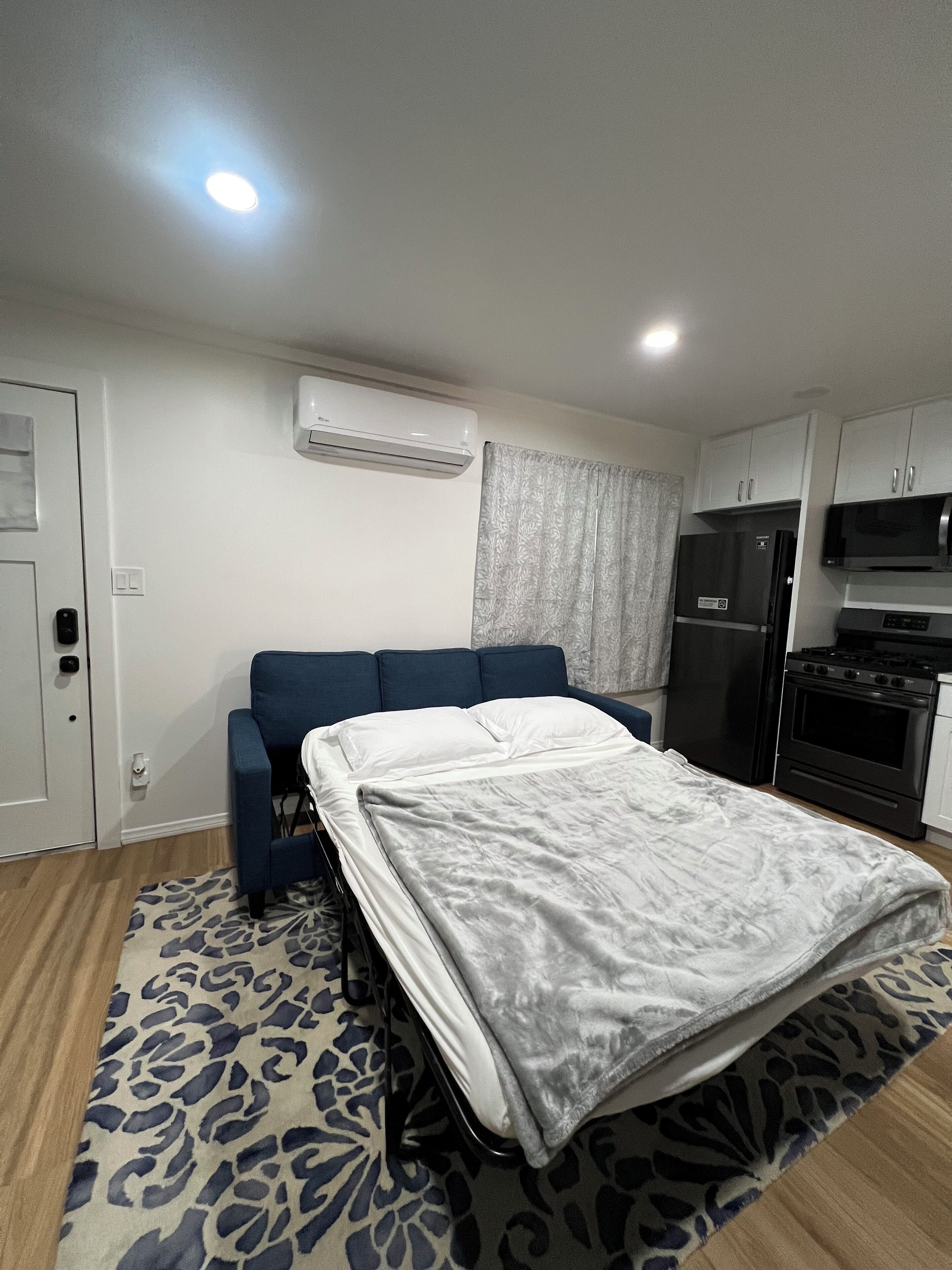 A compact room with a bed, a sofa, a kitchenette, and an air conditioning unit.