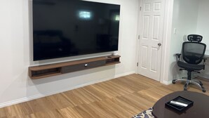 Una sala de estar moderna con un televisor de pantalla plana grande montado en la pared, un mueble de madera para equipo multimedia debajo, una silla de oficina negra y una pequeña mesa redonda con una bandeja.
