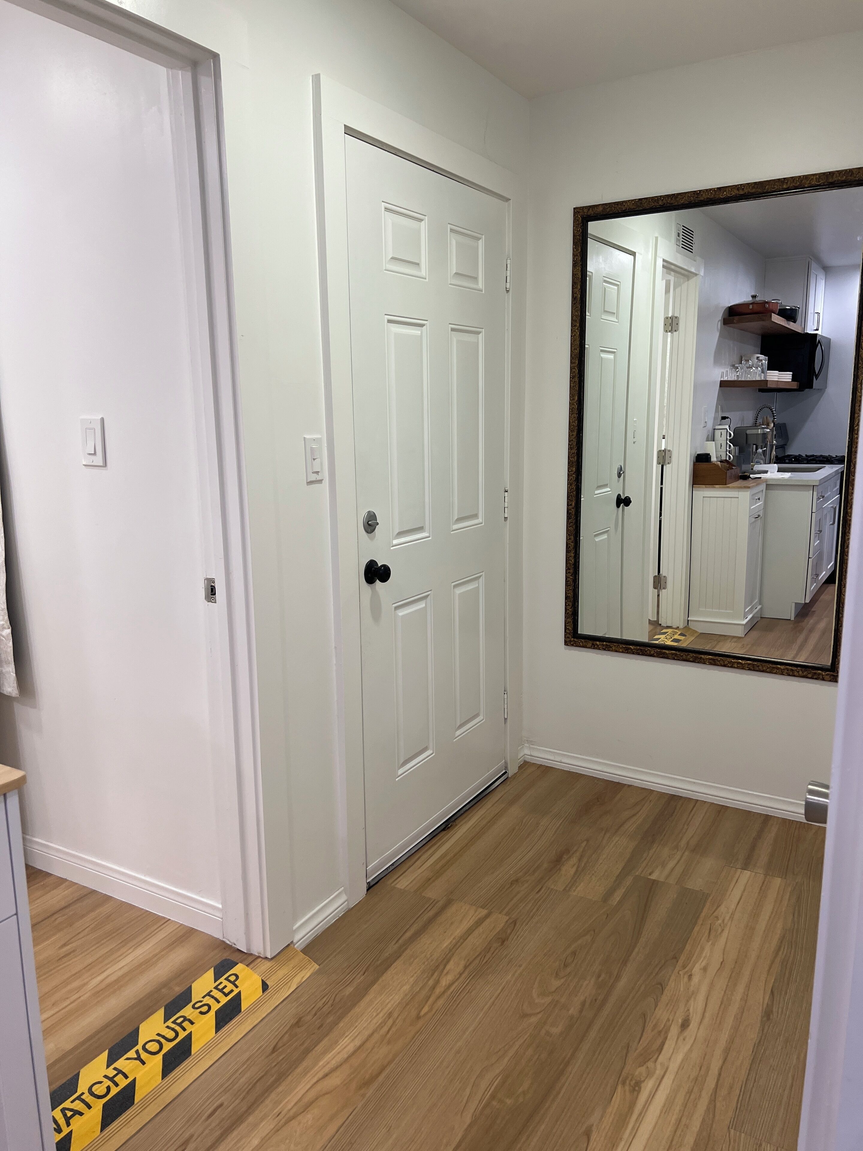 A hallway with white doors, a wooden floor, and a large mirror reflecting a kitchen area.