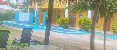 Un espacio al aire libre con una mesa y sillas bajo un techo de paja, rodeado de árboles y una piscina.
