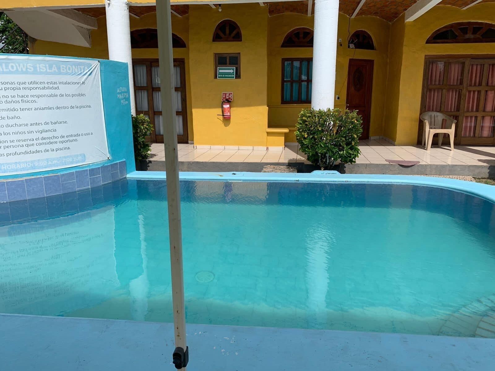 A swimming pool with a signboard, a covered area with chairs, and a yellow building with arched windows.