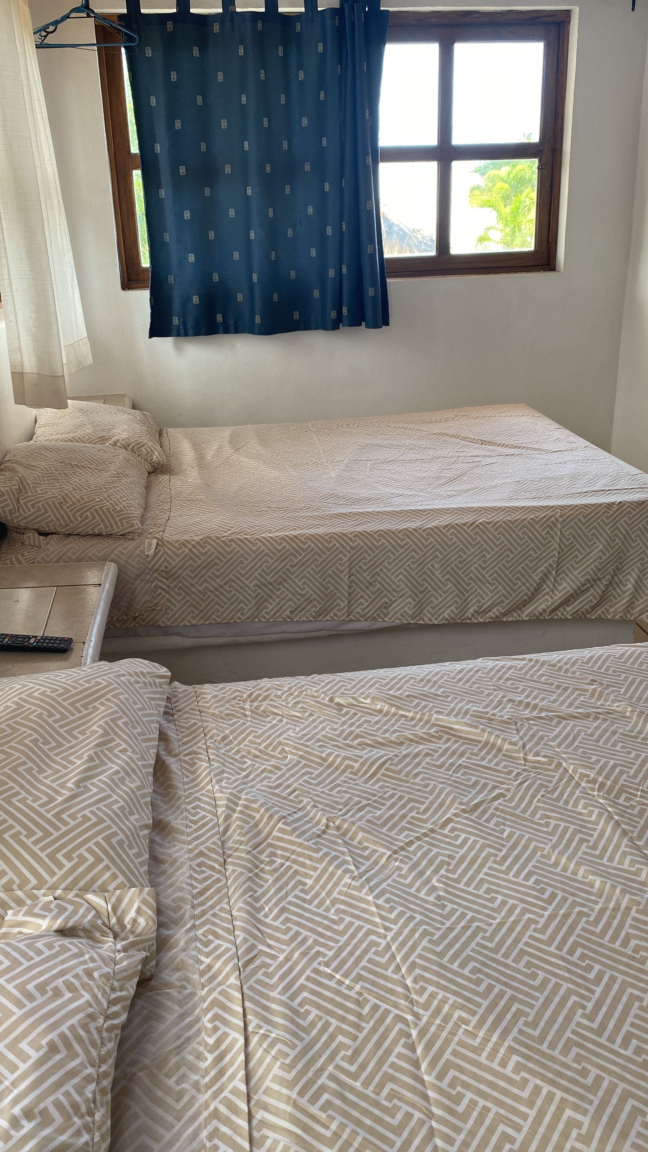 A room with two beds, a window with blue curtains, and a bed on the left with a remote control on it.
