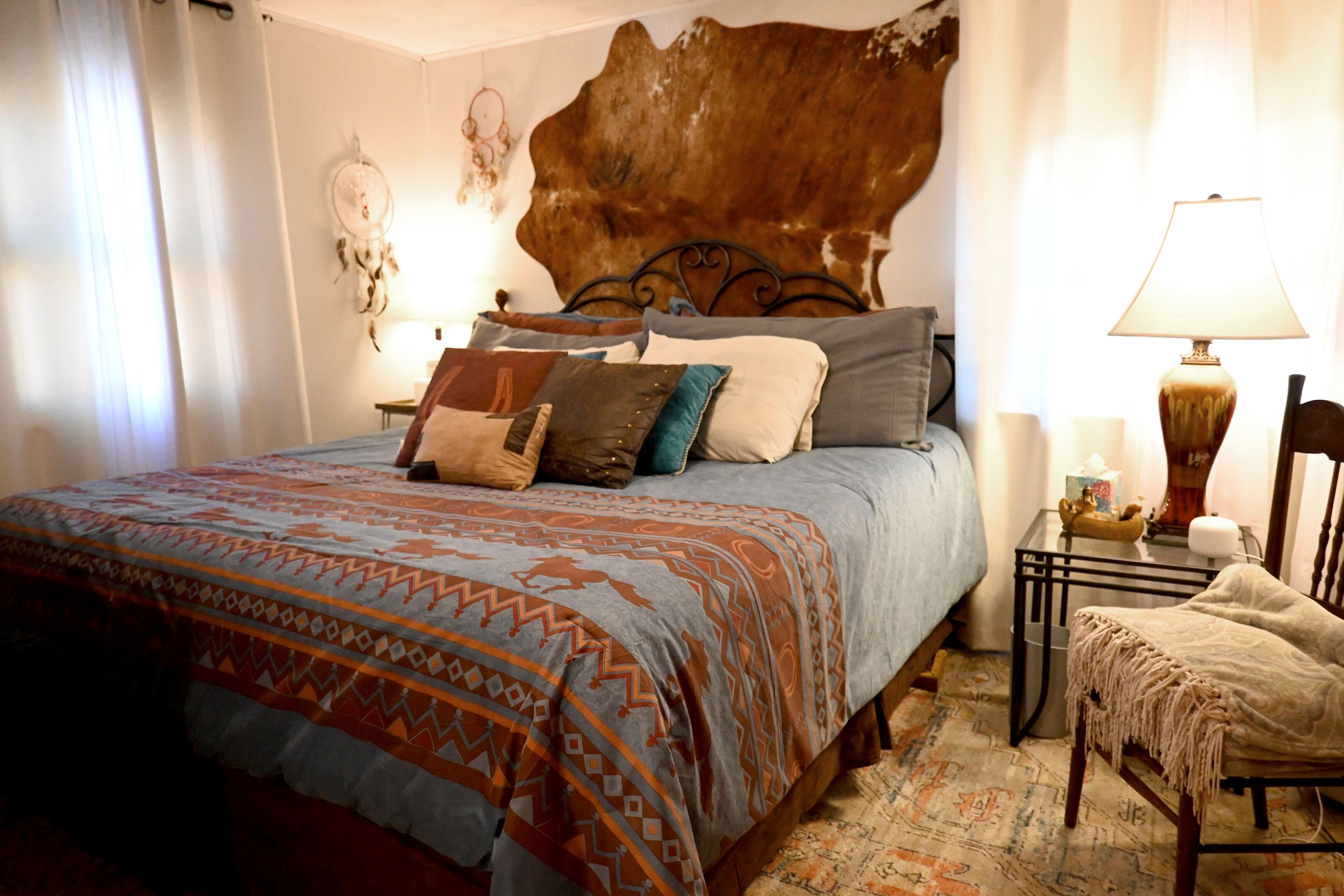 A bedroom with a large bed, a headboard, a bedside table with a lamp, a chair, and a patterned rug.