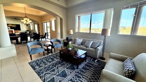 A spacious living room with a sofa, two armchairs, a coffee table, and a patterned rug. There is a dining area with a table and chairs, a kitchen in the background, and large windows offering a view of the outdoors.