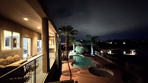 A nighttime view of a pool area with a staircase leading to a balcony, palm trees, and a cityscape in the background.