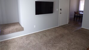 A room with a flat-screen TV mounted on the wall, carpeted flooring, a door, and a dining area in the background.