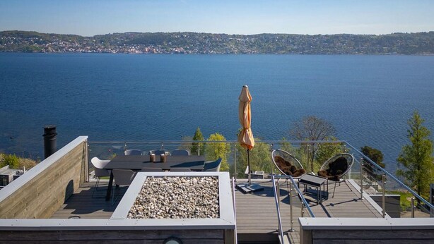 A deck with outdoor seating, a table, and an umbrella overlooking a lake with a distant town. - 4 bedroom nice home in Sætre (Sætre)