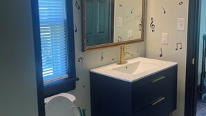 A bathroom with a white toilet, a dark blue sink with a gold faucet, and a mirror above the sink. There are three wall-mounted lights above the mirror. - Charming 2-bedroom cottage in wonderful Newburg with WiFi, AC (Newburg)
