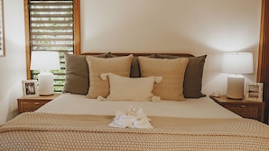 Una cama bien hecha con una colcha color crema, dos mesitas de noche con lámparas y una ventana con persianas.