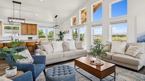 A spacious living room with a large sofa, a coffee table, and a dining area with wooden cabinets and a kitchen in the background. - Modern Lakehouse l Slps 34 l Courts + Chefs Kitchen l Bear Lake Haven (Saint Charles)