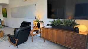 A modern living room with a large flat-screen TV, a black leather chair, a wooden coffee table, and a cabinet with decorative plants.