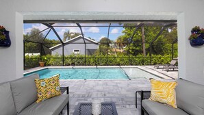Zona de piscina al aire libre cubierta con un sofá y cojines grises, una mesa y vistas a una casa y vegetación.
