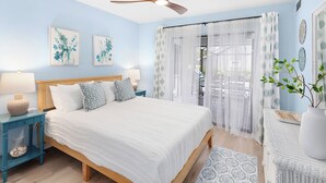 Un dormitorio con cama, dos mesitas de noche, ventilador de techo y ventana con cortinas traslúcidas.