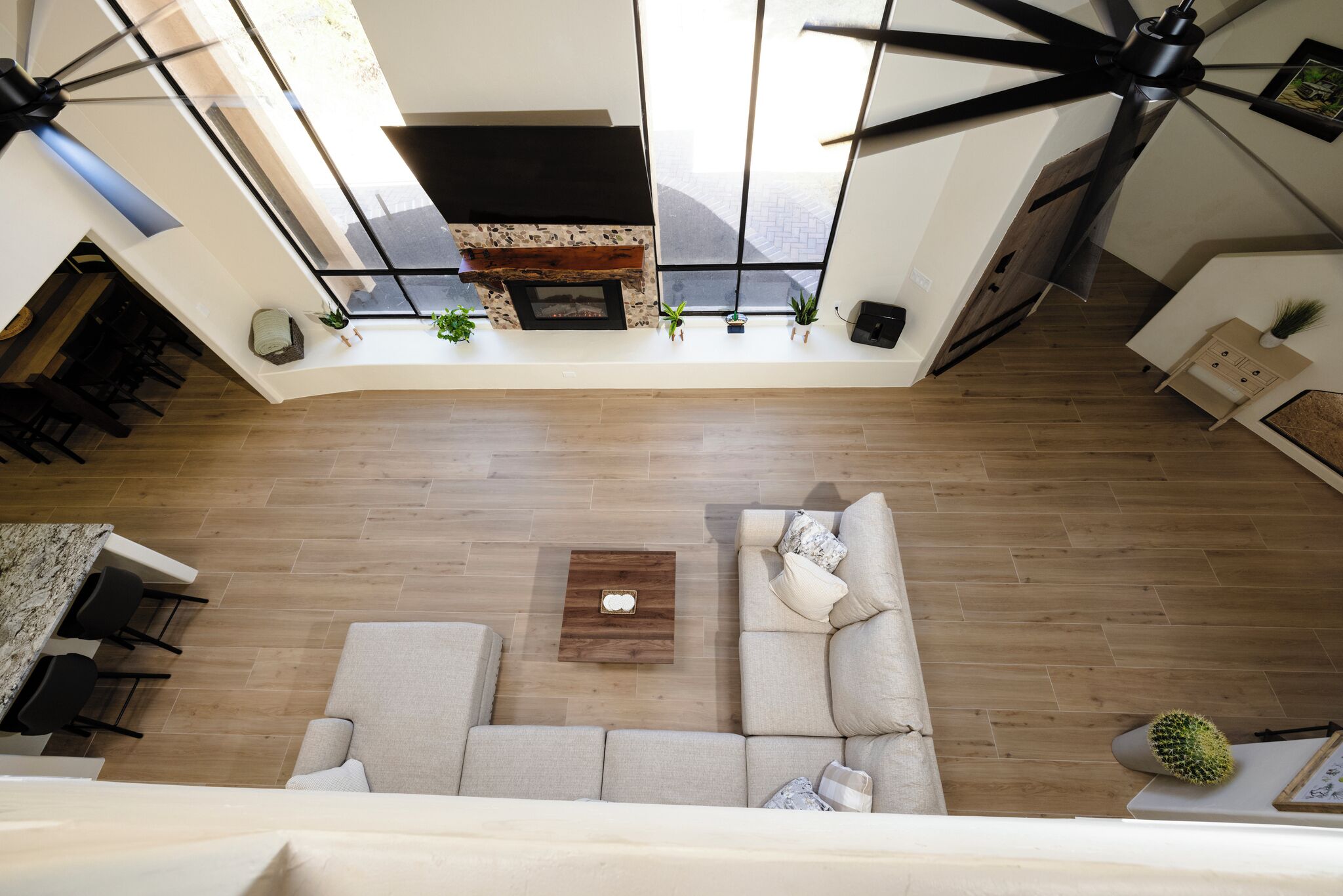 A modern living room with a sectional sofa, a coffee table, and a fireplace. There are ceiling fans, a dining area with chairs, and a kitchen visible.