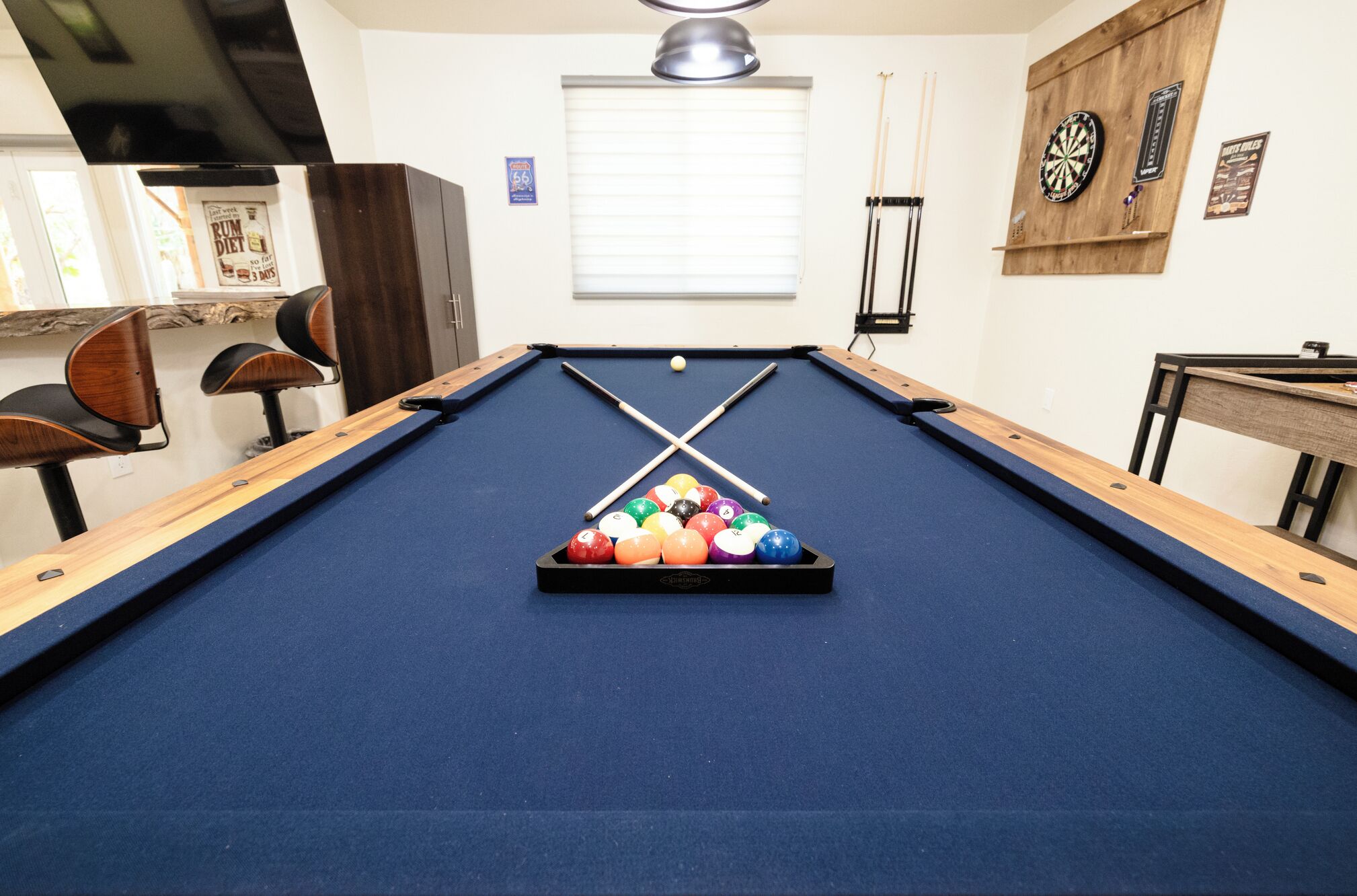 A pool table with a blue felt surface, surrounded by wooden chairs and a dartboard.