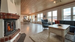 Ett rymligt vardagsrum med en öppen spis, en matplats och stora fönster med utsikt över omgivningen.