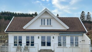 Ett vitt hus i två våningar med ett brunt tak täckt av tegelpannor, en veranda med stolar och en stenmur i bakgrunden.