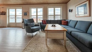 Ett vardagsrum med ett träpanelat tak, stora fönster, en grå soffa, ett träbord och en grå fåtölj.