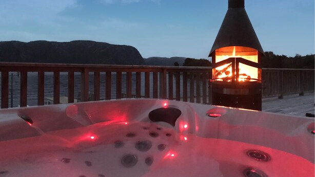 Ett bubbelbad med en eldstad, ett trädäck och en utsikt över vattnet och kullarna i bakgrunden.