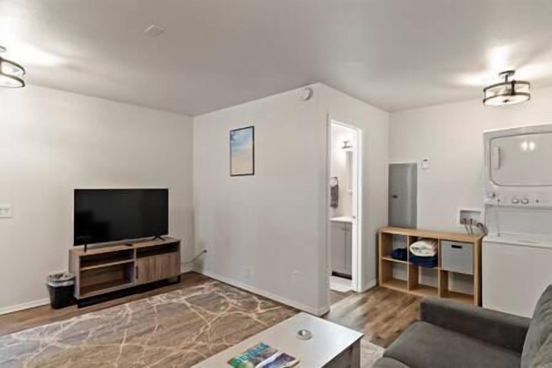 Ein kompakter Wohnbereich mit Flachbildfernseher, einem kleinen Couchtisch und einer Couch. Eine Tür führt ins Badezimmer, und es gibt einen weißen Schrank mit Regalen.